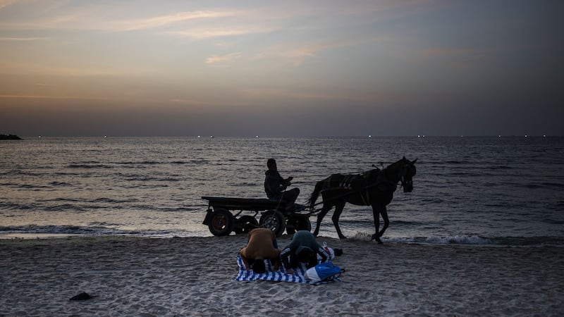 A man on a horse cart passes by two Palestinians pray before breaking their fast of a Ramadan Day on the beach in Gaza City, Sunday, May 2, 2021 (AP Photo/Khalil Hamra)&nbsp;
