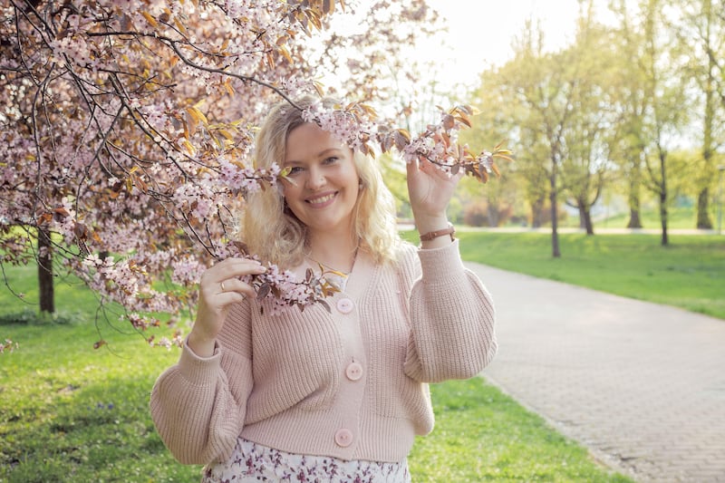 2J8CWCF Portrait of smiling beautiful blonde hair woman in pink knitted cardigan holding branches of blooming lilac on sunny day, nature background. Fashion