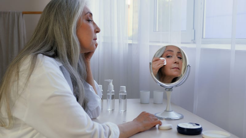 Middle aged woman sitting at a dressing table, looking in a mirror whilst removing make-up with a cotton wool pad