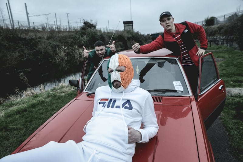 West Belfast rap trio Kneecap pose with a red 1980 Mazda 626