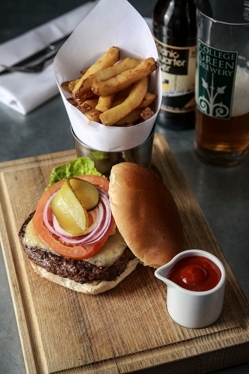 Image of an open burger - showing the patty, cheese, lettuce, red onion and gherkin in it. There also some chips in a small silver bucket beside the plate and a small ramekin of tomato sauce.