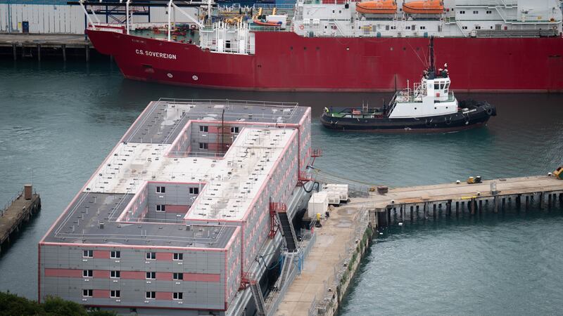 A tug boat passing the Bibby Stockholm accommodation barge at Portland Port in Dorset
