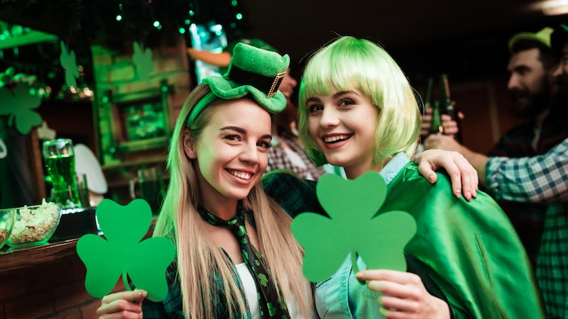 Two girls in a wig and a cap are photographed in a bar. They celebrate St. Patrick's Day. They are having fun. One girl is holding a clover.