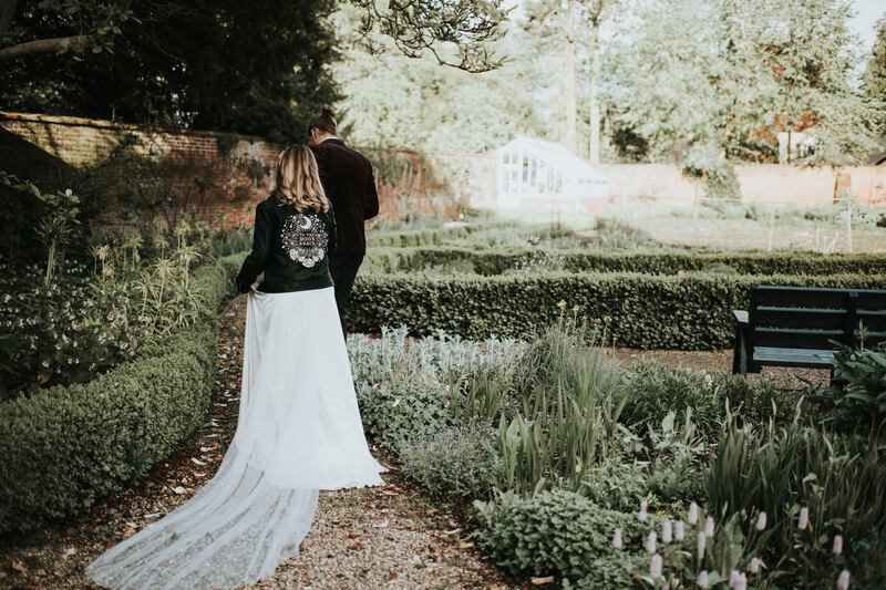 The backs of a bride and groom walking in a garden. Bride is wearing a black leather jacket with the moon and stars embroidered on it and the words "to the moon and back".