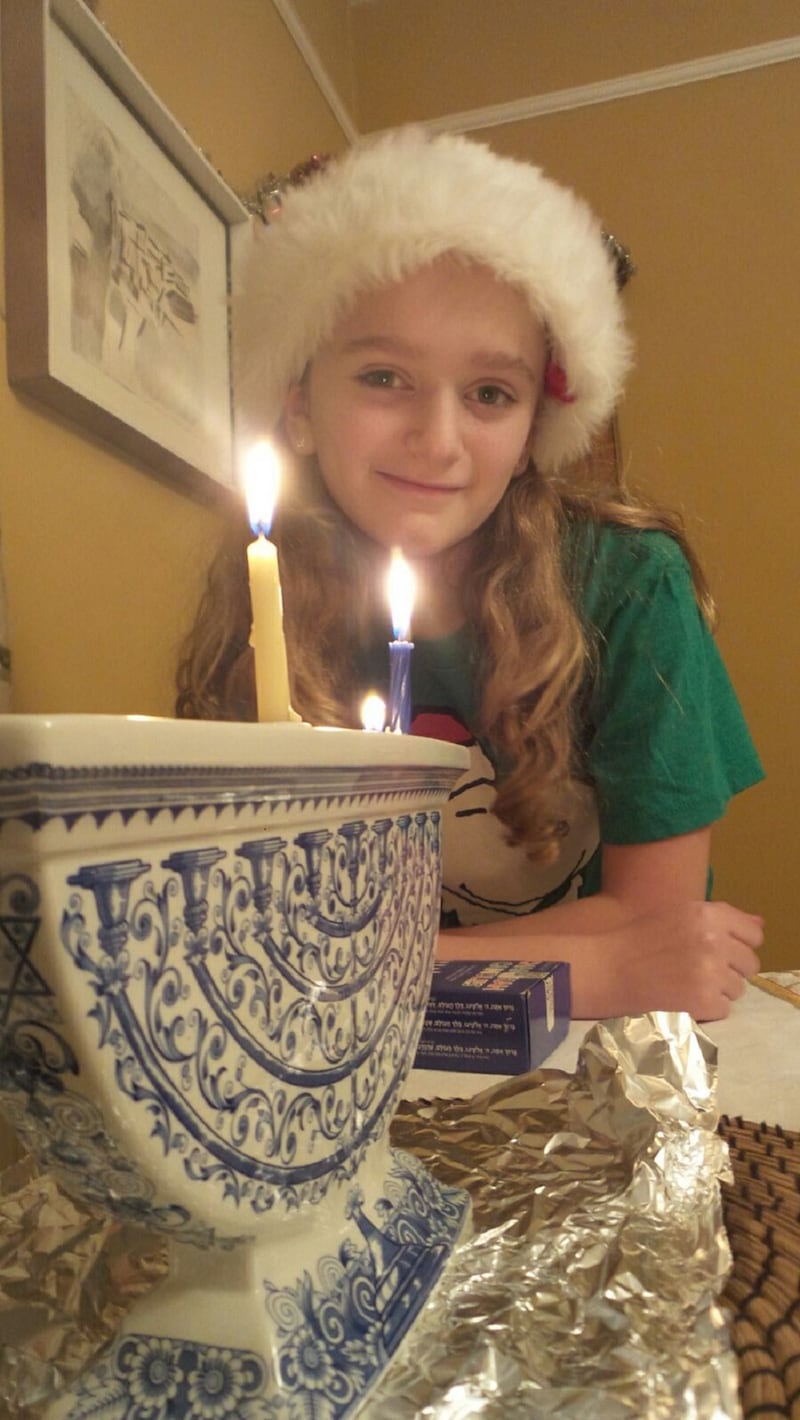 Bea Baldwin as a young child wearing a Santa hat and standing in front of a menorah