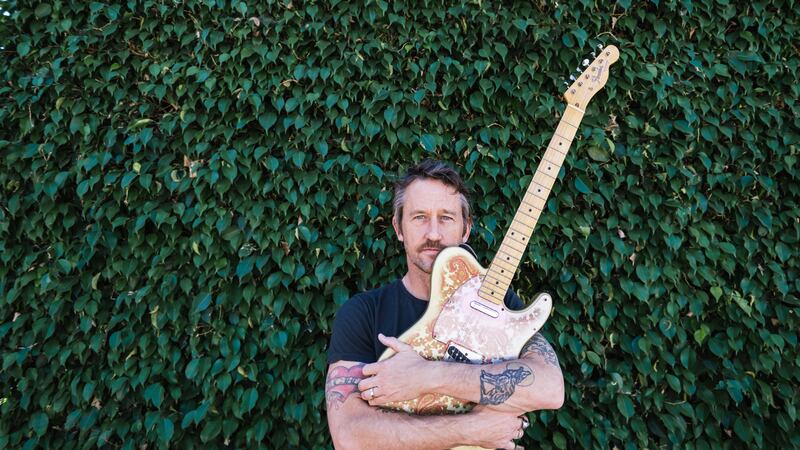 A phot of Foo Fighters' guitarist and solo artist Chris Shiflett, with his signature Fender Telecaster guitar cradled in his arms, standing in front of a hedge