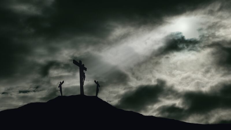 A depiction of the crucifixion of Jesus Christ on a cross with 2 other robbers nearby on Calvary. The sky is darkened with rays of light breaking through the clouds onto the cross for drama. Concept of the death of Jesus on Good Friday and His resurrection on Easter Sunday. Horizontal orientation with copy space.