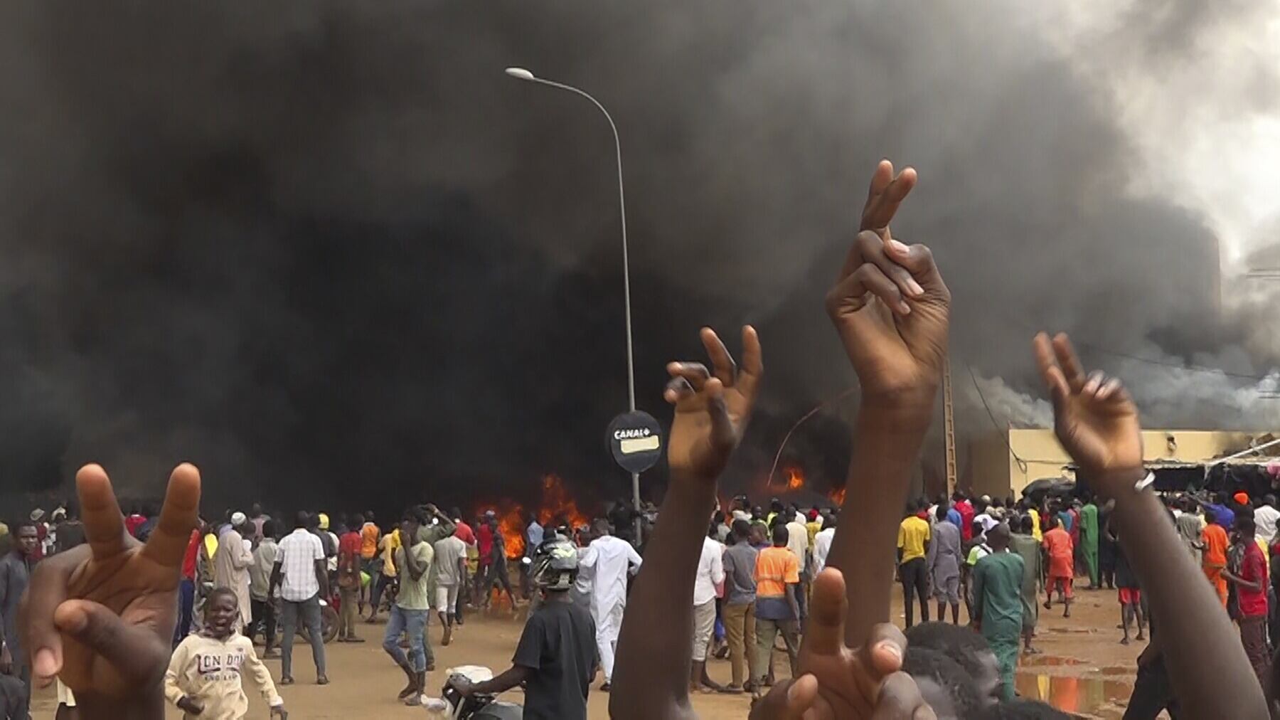 With the headquarters of the ruling party burning in the back, supporters of mutinous soldiers demonstrate in Niamey (Fatahoulaye Hassane Midou/AP)
