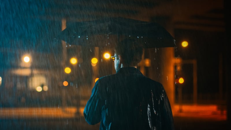 Backshot of a Young Caucasian Shorthaired Man, Wearing a Jeans Coat Walking in the Rain Under an Umbrella. He's Walking at Night in the City.