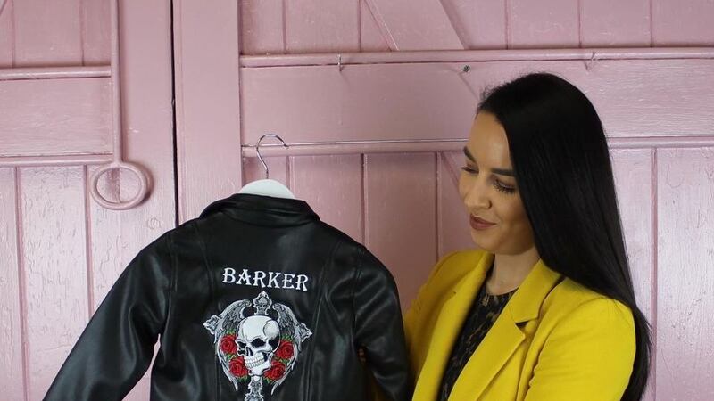 Niamh in a yellow blazer, holding the baby sized leather jacket. There is a skull and roses embroidered on the jacket.