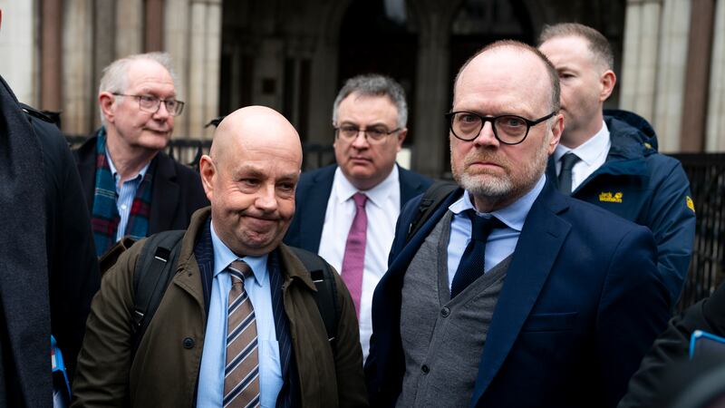 Barry McCaffrey on the left is dressed in a suit and tie with a knapsack on his back. He stands beside Trevor Birney who is wearing a navy suit jacket, grey waistcoat and dark tie. They are standing outside the Royal Courts of Justice in London with supporters gathered behind them