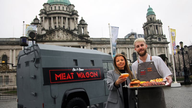 A man and woman holding a tray of food and drink in front of an old RUC land rover with the Meat Wagon Signage parked in front of Belfast City Hall.