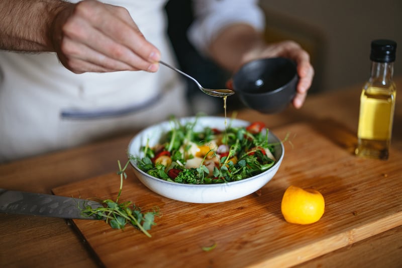 High angle view of an anonymous chef in apron pouring a teaspoon of salad oil into a fresh salad with micro green herbs at the kitchen table.