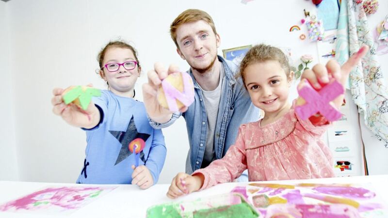 Ulster University student facilitator Conor McClure helps sisters Eimear and Cara Gallagher print potato designs on a canvas bag 