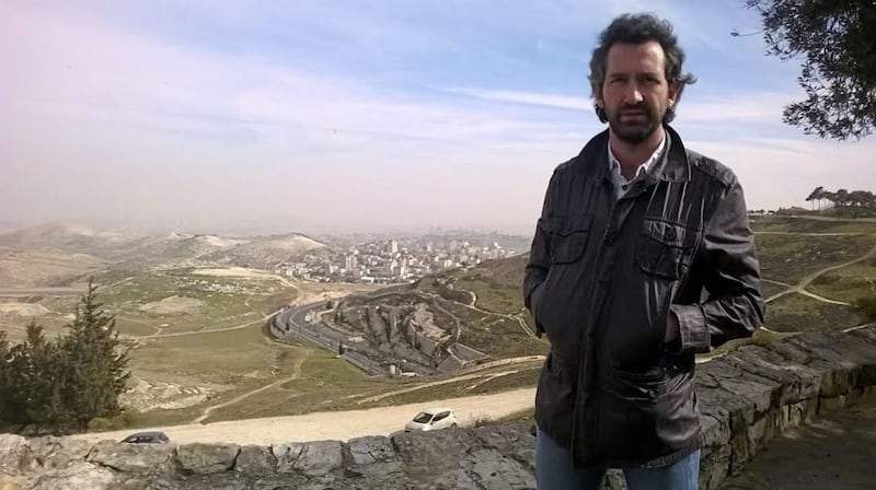 Dr Conn Mac Gabhann; behind him is a view from Jerusalem of a Palestinian village and an Israeli settlement 